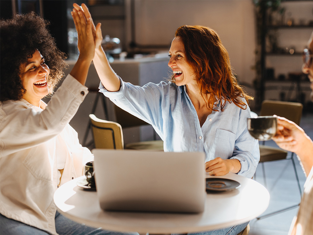 Primo piano di una mentor e una mentee middle manager che celebrano un successo dandosi il cinque, davanti a un laptop.​