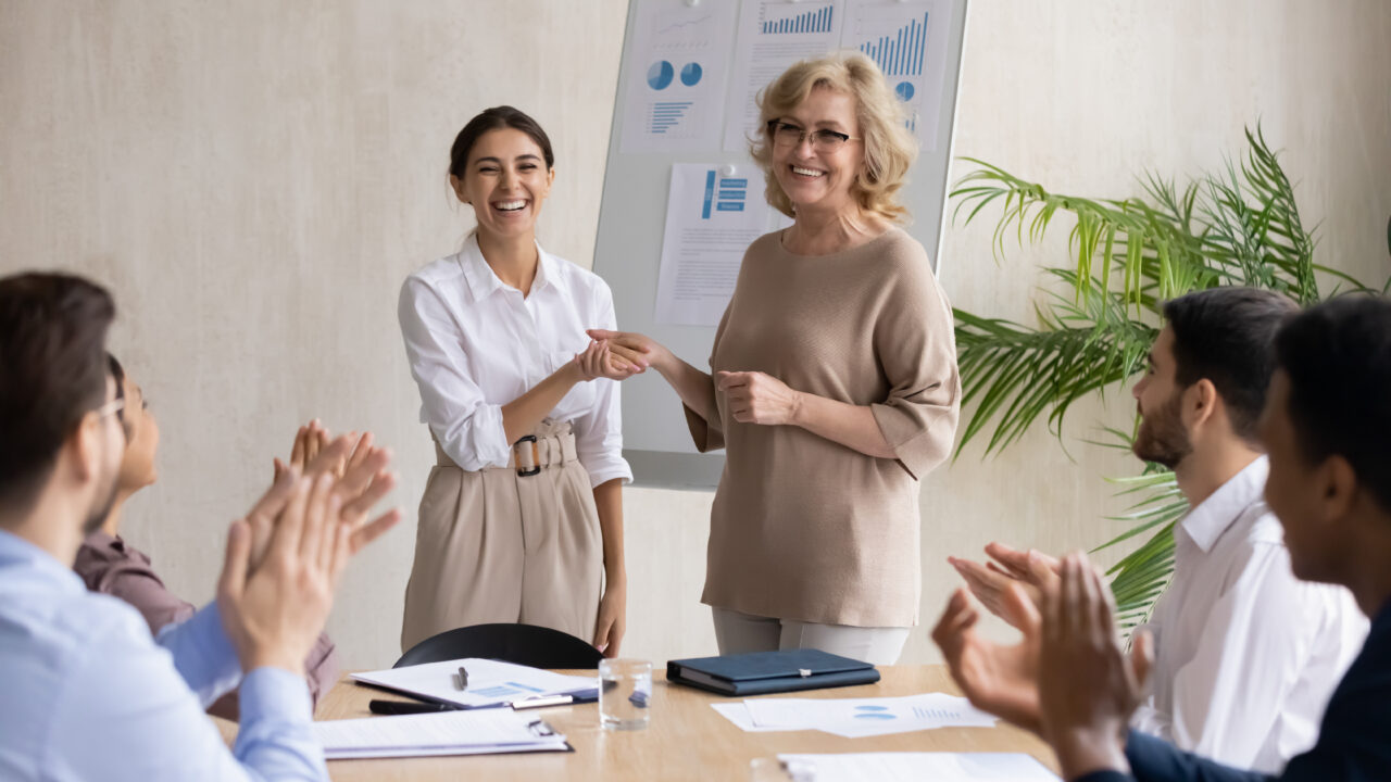 Due donne (una di età alta, una di età bassa) si stringono la mano sorridenti davanti ai colleghi seduti a un tavolo in ufficio. Dietro di loro il poster di una presentazione.