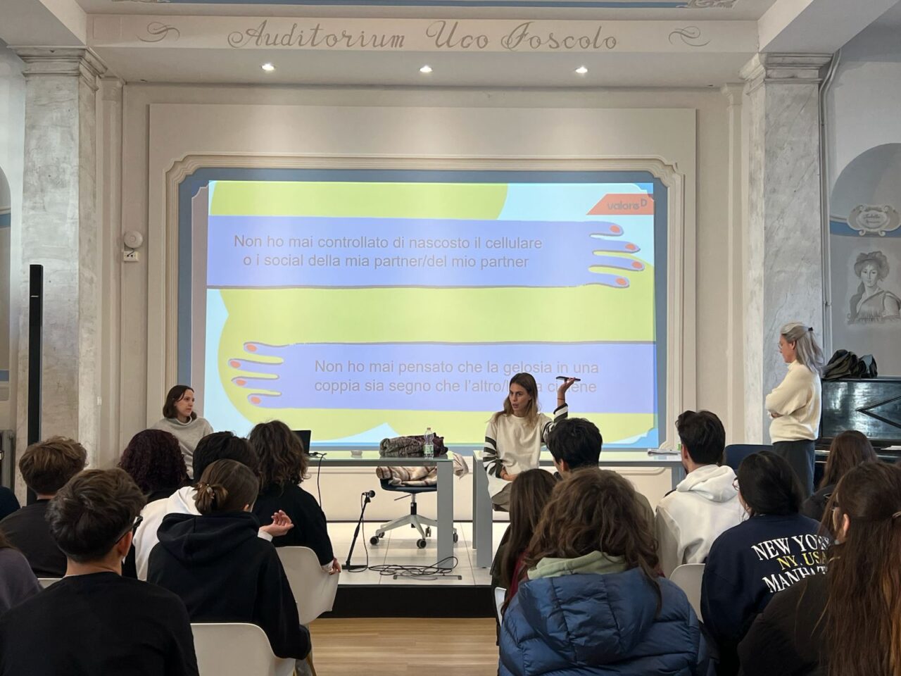 Immagine dell'incontro "Scomodiamoci a Scuola". Carolina de Castiglioni e Chiara Gregori parlano a una platea di ragazze e ragazzi. Dietro di loro una slide del gioco "Non ho mai".