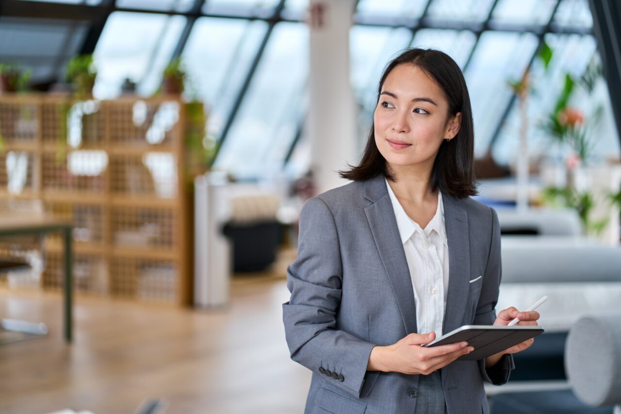 Donna di origini asiatiche con tablet in mano e sguardo rivolto alla sua destra. Indossa un tailleur elegante e si trova in una sala d'ufficio senza altre persone.